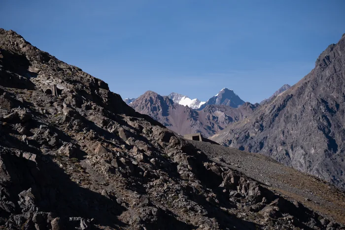 Rugged Andes mountain range with snow-capped peaks visible in the distance beyond rocky foreground slopes under clear blue sky
