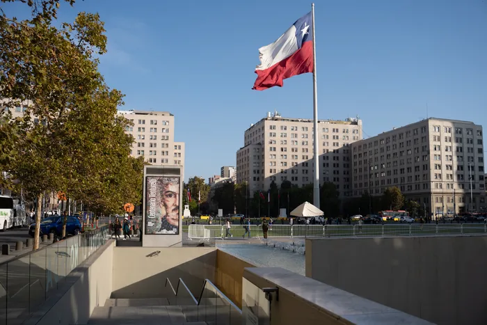 The Chilean flag waving over Plaza de Armas in Santiago, with classical buildings and a metro entrance in the foreground.