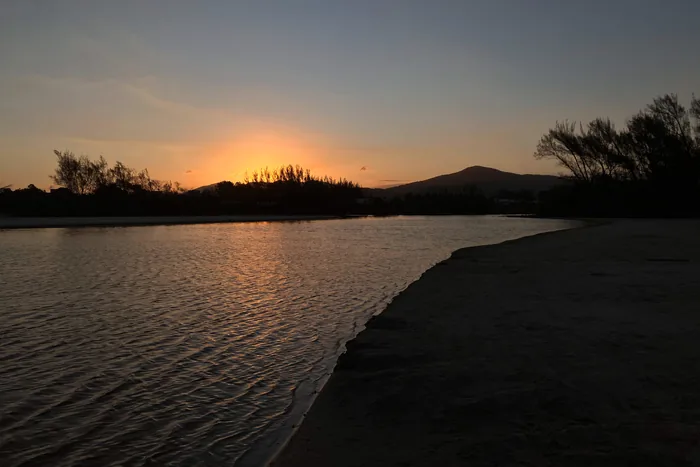 Tranquil lagoon at sunset with orange glow on the horizon, silhouetted trees and mountains, and golden reflections on calm water