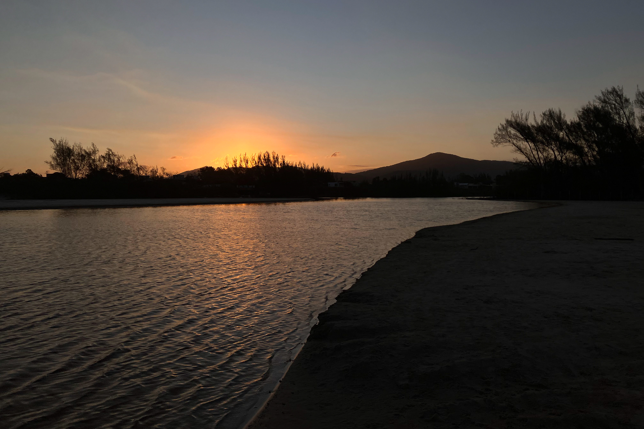 Tranquil lagoon at sunset with orange glow on the horizon, silhouetted trees and mountains, and golden reflections on calm water