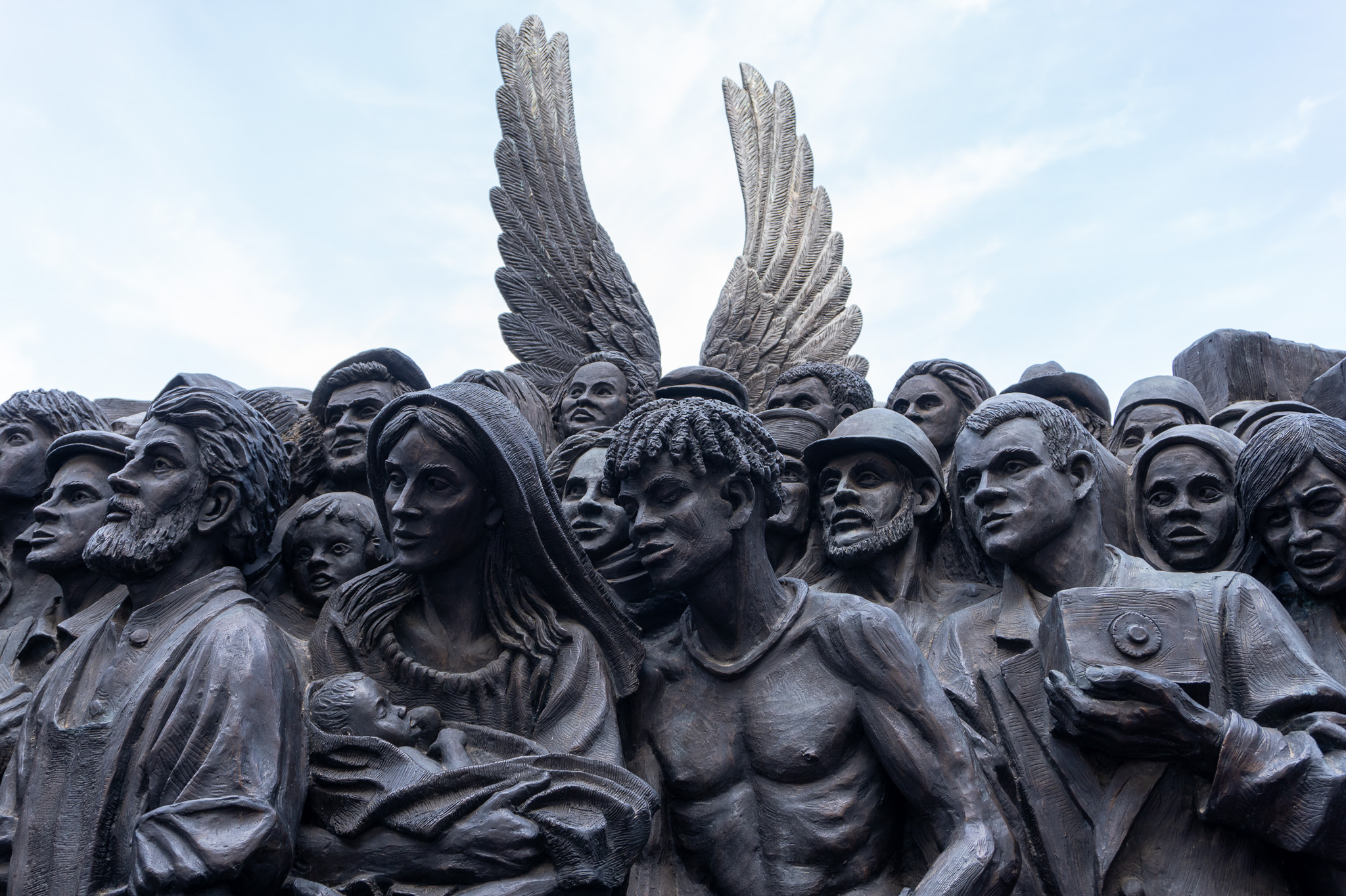 A large bronze sculpture depicting a dense crowd of diverse people, with a pair of angel wings rising above them against a bright sky.