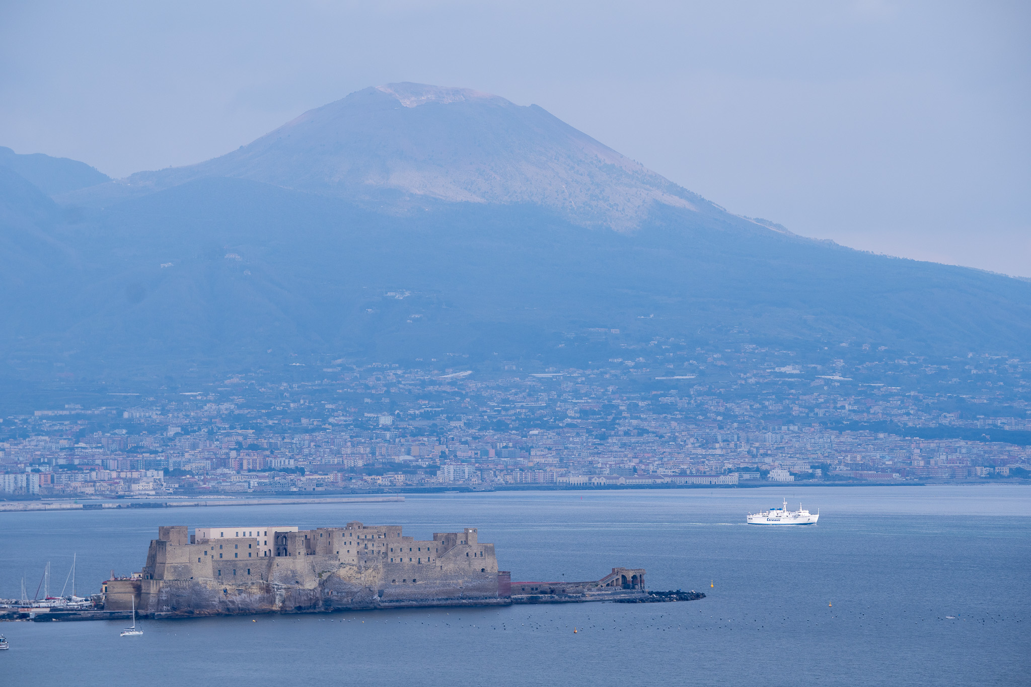 Castel dell'Ovo on a rocky islet in the Bay of Naples, with Mount Vesuvius looming in the misty background and a ferry crossing the water.