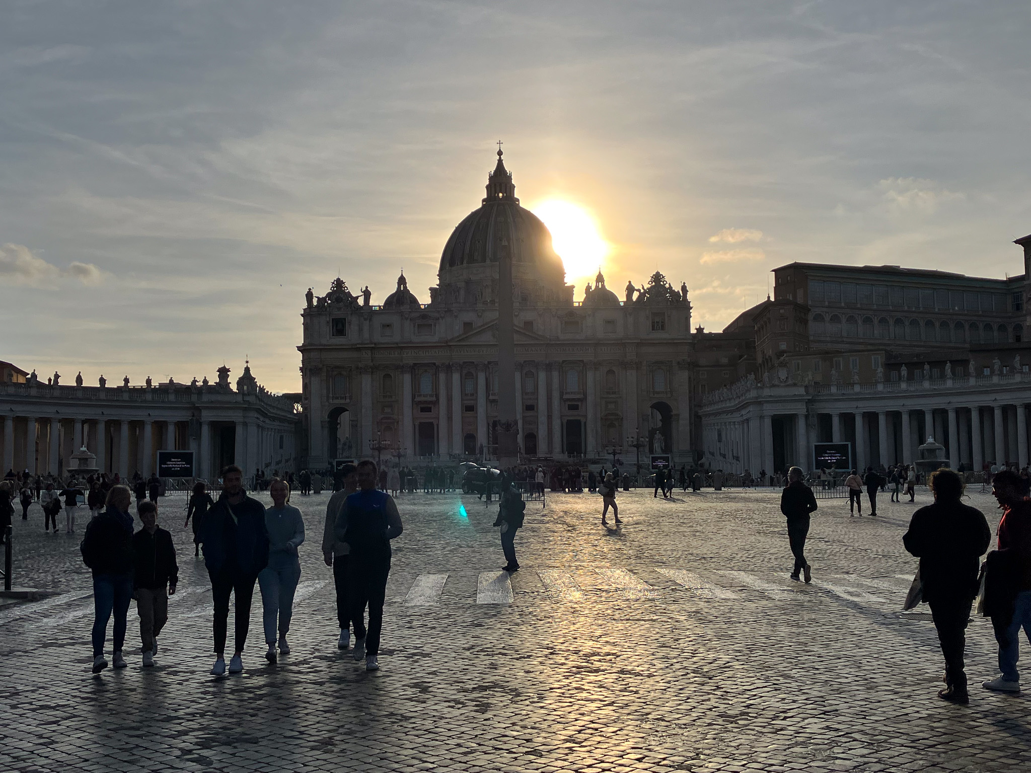 St. Peter's Basilica at Vatican City seen from St. Peter's Square at sunset, with the sun behind the dome casting a golden glow and silhouettes of visitors walking across the cobblestones.