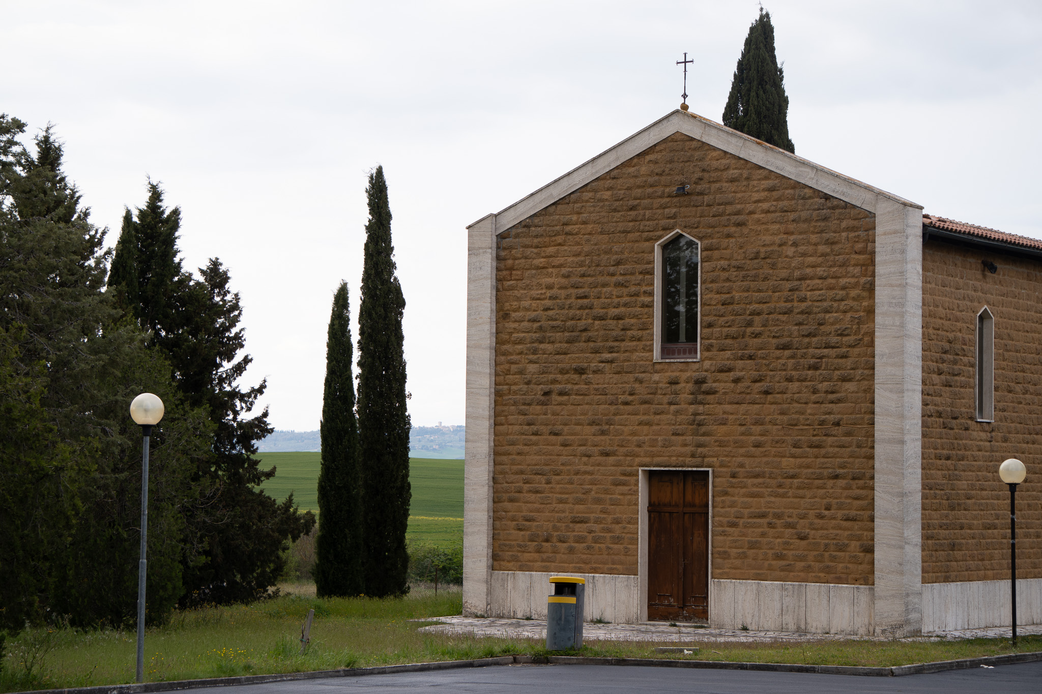 A simple stone church with a cross on the rooftop, flanked by tall dark cypress trees and surrounded by a flat green field under an overcast sky.