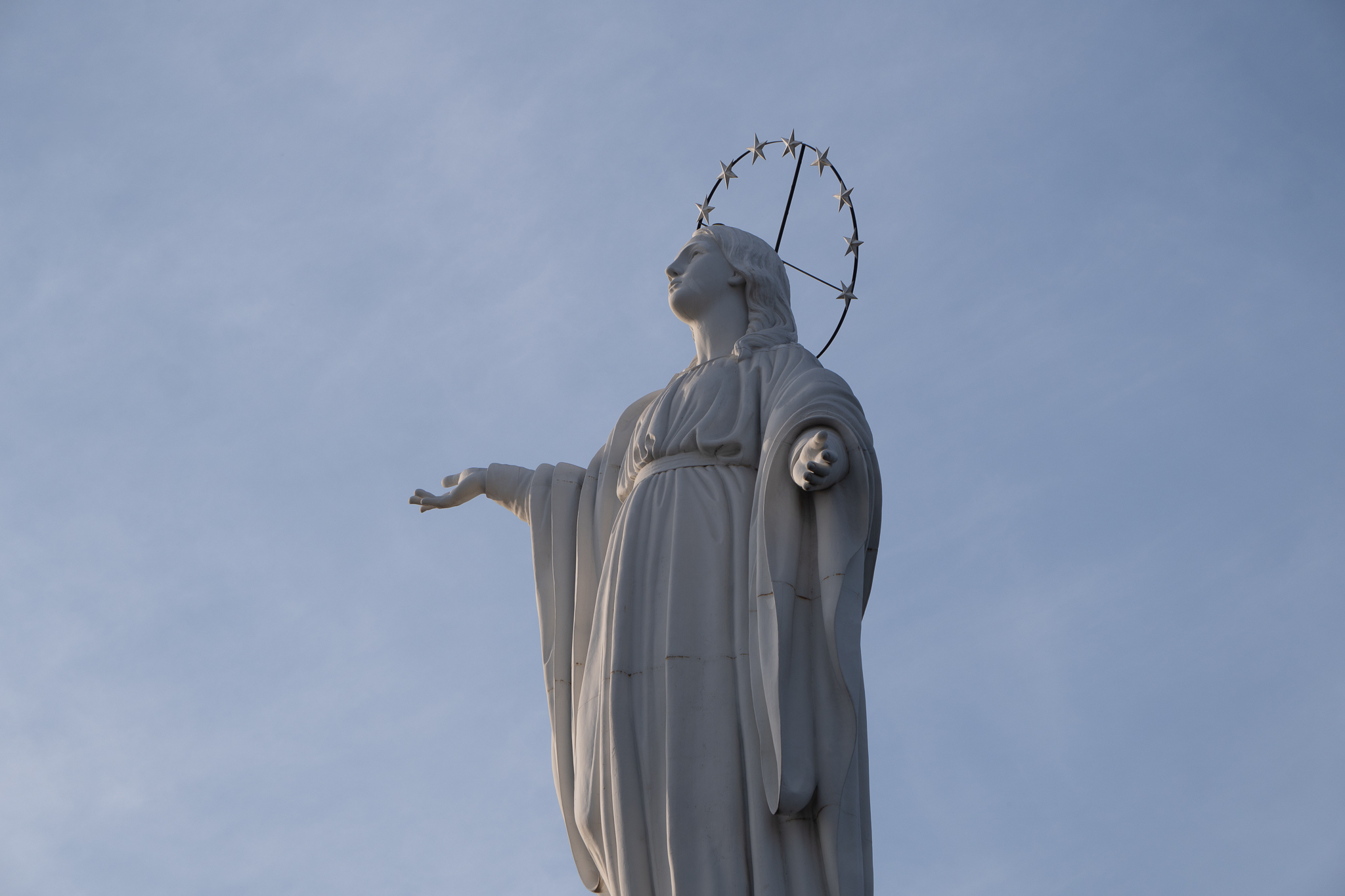 White statue of the Virgin Mary atop Cerro San Cristóbal with outstretched arm and star-crowned halo against clear blue sky