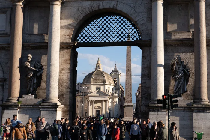 A crowd of people seen through the large arch of Porta del Popolo in Rome, with the twin churches and an Egyptian obelisk visible in Piazza del Popolo beyond.