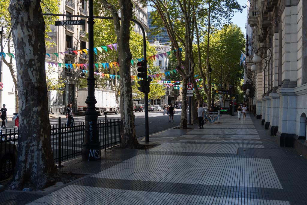 A wide tree-lined pedestrian boulevard in downtown Buenos Aires with colonial-style buildings, colorful bunting flags strung between the trees, and the Bolivar street sign visible in the foreground.