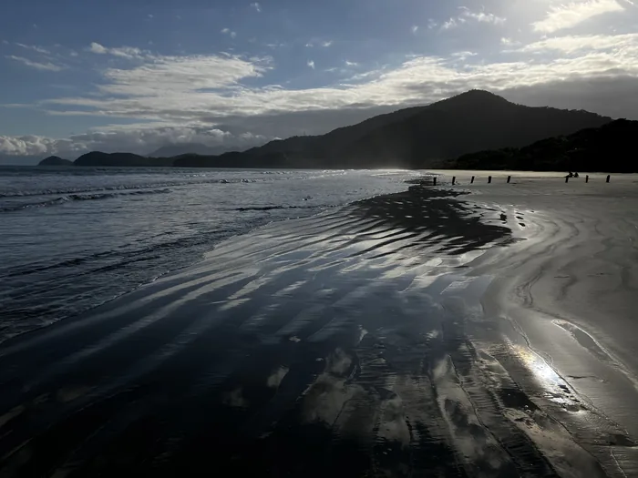 Fazenda Beach at low tide with sunlight reflecting across wet sand and forested mountains along the coast
