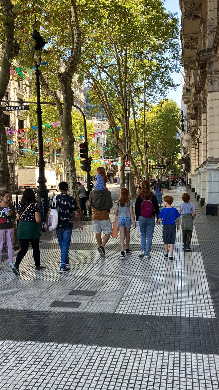 People walking along a sun-dappled pedestrian street in Buenos Aires lined with plane trees and colorful bunting flags, with the Bolivar street sign visible.